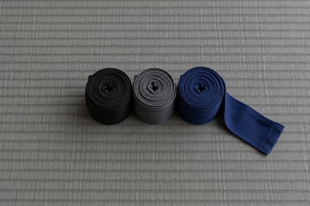 Trois bandes de boxe roulées, noire, grise et bleu marine, alignées sur un tatami gris